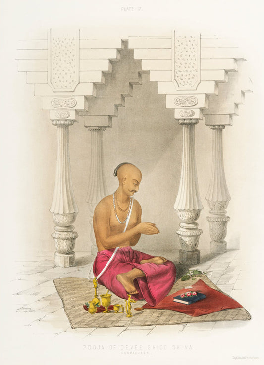 A priest in a rose-coloured garment sits between carved pillars, with a small arrangement of offerings at his feet.